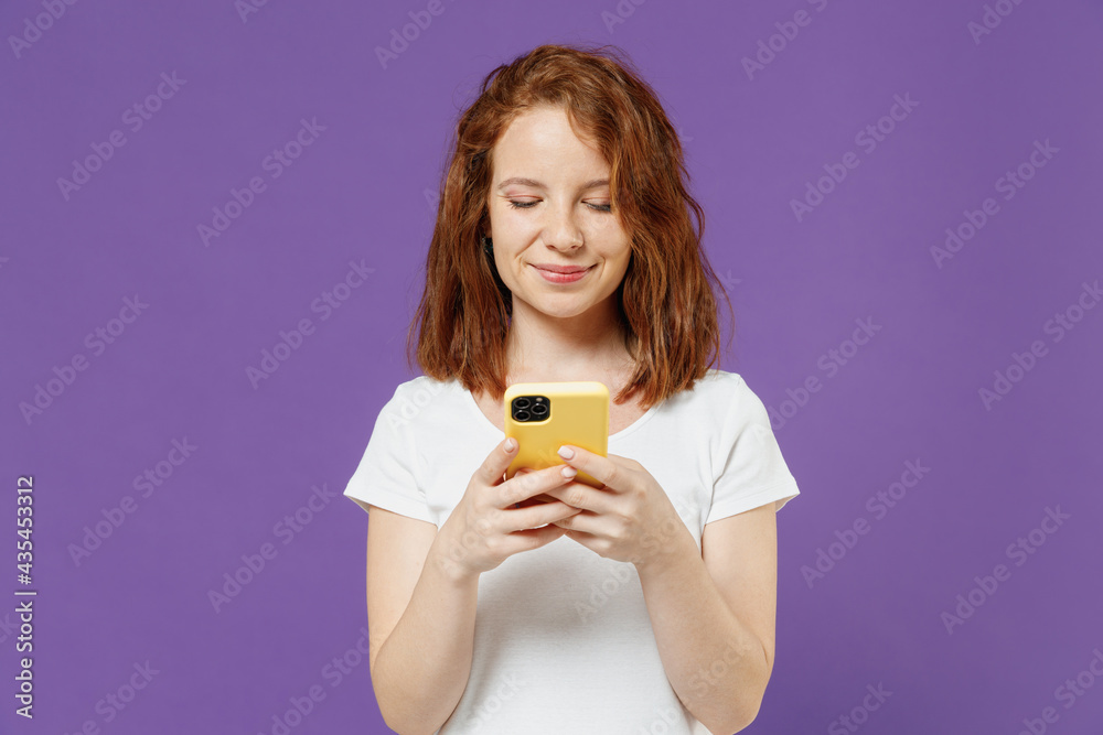 Young smiling happy satisfied fun redhead woman 20s in white basic casual t-shirt holding using mobile cell phone chat online browsing search isolated on dark violet color background studio portrait Young smiling happy satisfied fun redhead woman 20s in white basic casual t-shirt holding using mobile cell phone chat online browsing search isolated on dark violet color background studio portrait
