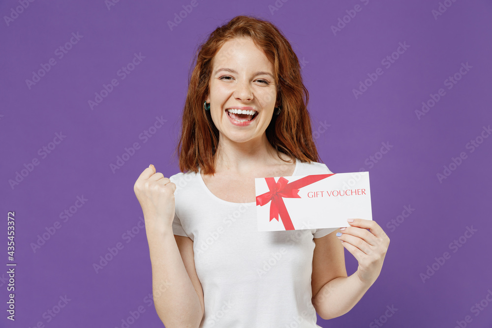 Young happy redhead caucasian woman 20s in white basic blank print design t-shirt holding gift voucher flyer mock up do winner gesture clench fist isolated on dark violet background studio portrait Young happy redhead caucasian woman 20s in white basic blank print design t-shirt holding gift voucher flyer mock up do winner gesture clench fist isolated on dark violet background studio portrait
