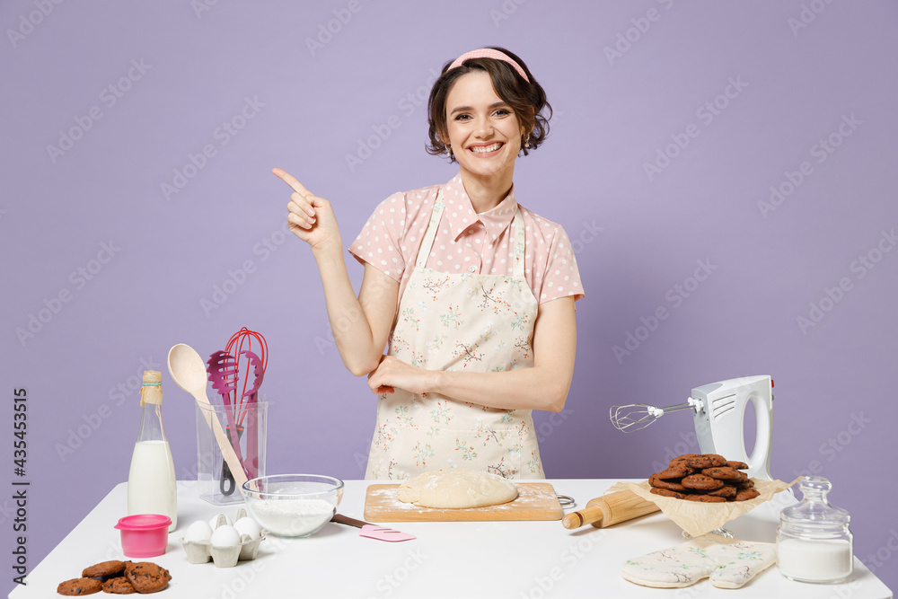 Young fun housewife housekeeper chef cook baker woman in pink apron work at table kitchenware point index finger aside on copy space isolated on pastel violet background. Process cooking food concept. Young fun housewife housekeeper chef cook baker woman in pink apron work at table kitchenware point index finger aside on copy space isolated on pastel violet background. Process cooking food concept.