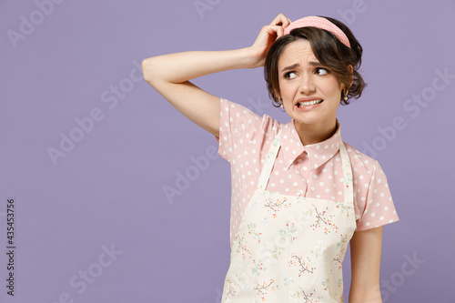 Young puzzled displeased troubled sad housewife housekeeper chef cook baker woman wearing pink apron scratch head look aside isolated on pastel violet background studio portrait. Cooking food concept