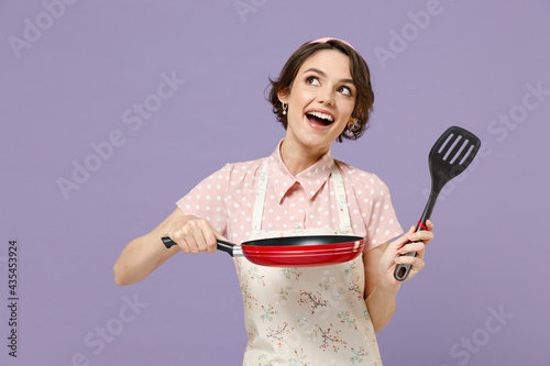 Young happy pensive housewife housekeeper chef cook baker woman in pink apron hold show red frying pan spatula look aside overhead isolated on pastel violet background Cooking food process concept photo