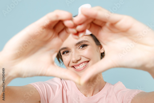 Young woman in pajamas jam sleep eye mask rest relaxing at home showing shape heart with hands look through heart-shape sign isolated on pastel blue background studio. Good mood night bedtime concept.