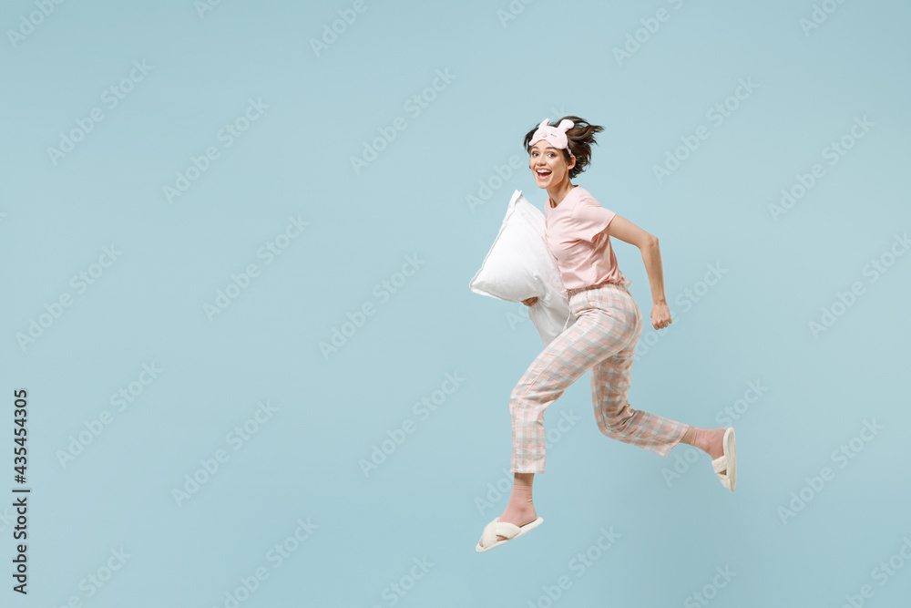 Full length side view young happy excited woman in pajamas jam sleep eye mask rest relaxing at home jump high with pillow run isolated on pastel blue background studio Good mood night bedtime concept Full length side view young happy excited woman in pajamas jam sleep eye mask rest relaxing at home jump high with pillow run isolated on pastel blue background studio Good mood night bedtime concept