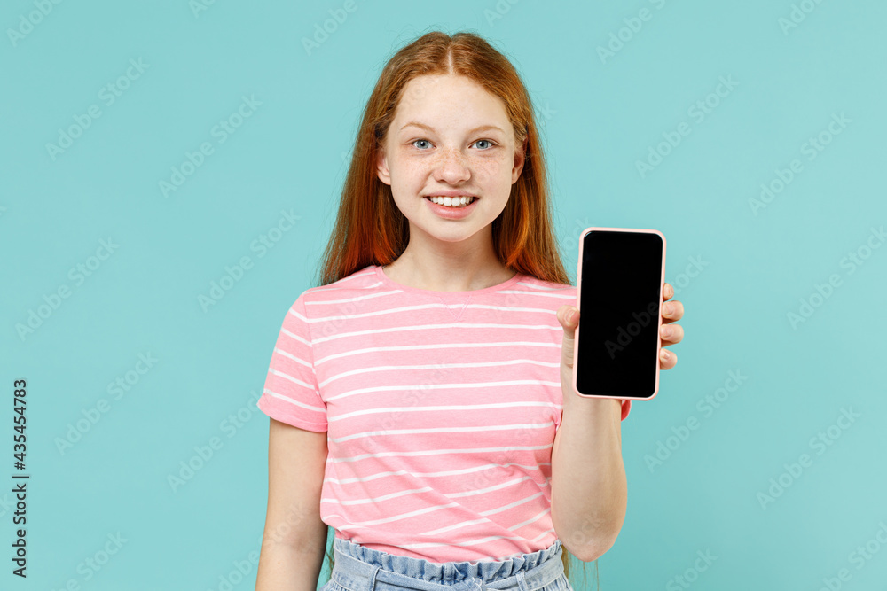 Little redhead kid girl 12-13 years old in pink striped t-shirt hold mobile cell phone with blank screen workspace area isolated on pastel blue background studio. Children lifestyle childhood concept Little redhead kid girl 12-13 years old in pink striped t-shirt hold mobile cell phone with blank screen workspace area isolated on pastel blue background studio. Children lifestyle childhood concept