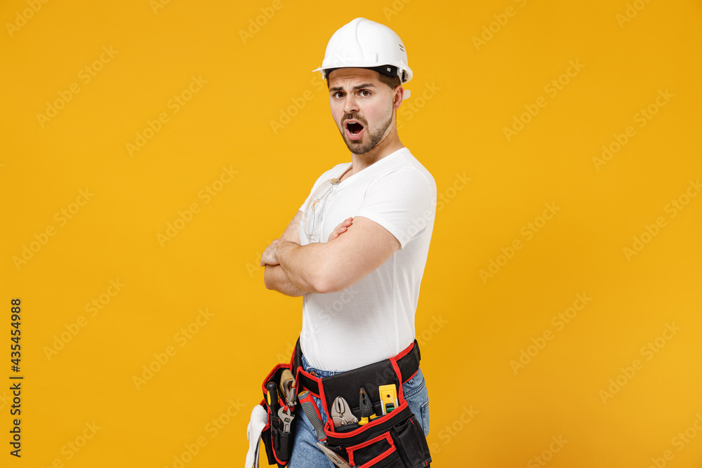 SIde view young employee handyman man in protective helmet hardhat hold hands crossed folded isolated on yellow background. Instruments accessories for renovation apartment room. Repair home concept. SIde view young employee handyman man in protective helmet hardhat hold hands crossed folded isolated on yellow background. Instruments accessories for renovation apartment room. Repair home concept.