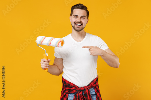 Young fun smiling employee handyman man wear t-shirt point index finger on paint roller isolated on yellow background studio. Instruments accessories for renovation apartment room Repair home concept. photo