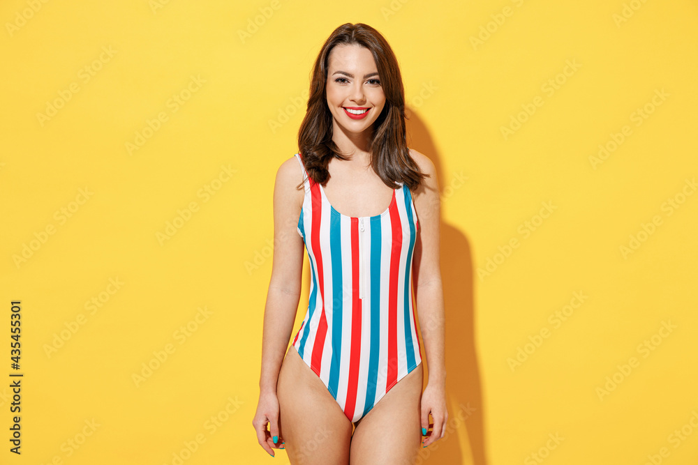 Happy fun smiling young sexy woman slim body wear striped red blue one-piece swimsuit looking camera isolated on vivid yellow color wall background studio. Summer hotel pool sea rest sun tan concept. Happy fun smiling young sexy woman slim body wear striped red blue one-piece swimsuit looking camera isolated on vivid yellow color wall background studio. Summer hotel pool sea rest sun tan concept.