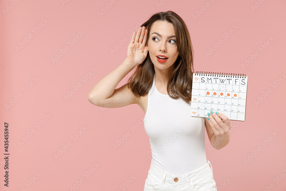 Young woman in white t-shirt hold female periods calendar checking menstruation days try to hear you overhear listening intently isolated on pink background Medical healthcare gynecological concept. Young woman in white t-shirt hold female periods calendar checking menstruation days try to hear you overhear listening intently isolated on pink background Medical healthcare gynecological concept.