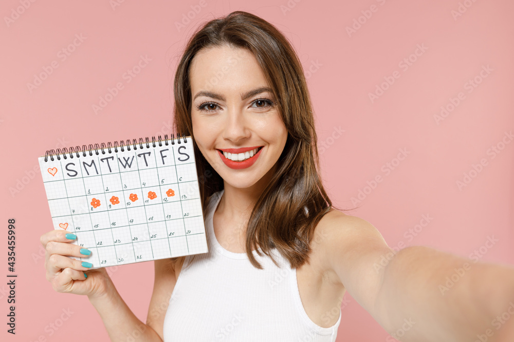 Close up happy young woman in white clothes hold female periods calendar checking menstruation days do selfie shot on mobile phone isolated on pink background Medical healthcare gynecological concept Close up happy young woman in white clothes hold female periods calendar checking menstruation days do selfie shot on mobile phone isolated on pink background Medical healthcare gynecological concept