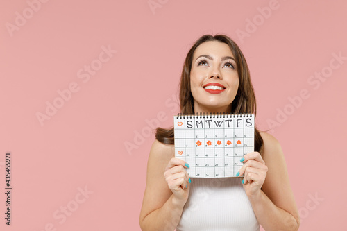 Young happy woman in white clothes hold in hand female periods calendar for checking menstruation days look overhead isolated on pastel pink background studio Medical healthcare gynecological concept