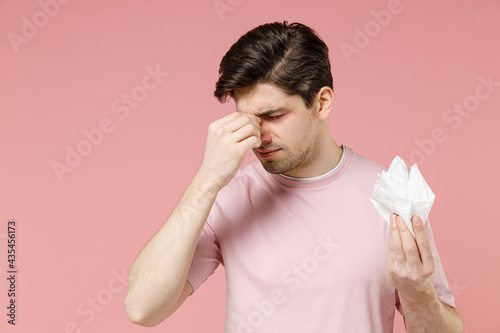 Sick ill allergic man has red eyes put hand on runny inflammation nose suffer from allergy symptoms hold paper napkin reacts on environmental allergen isolated on pastel pink color background studio. photo