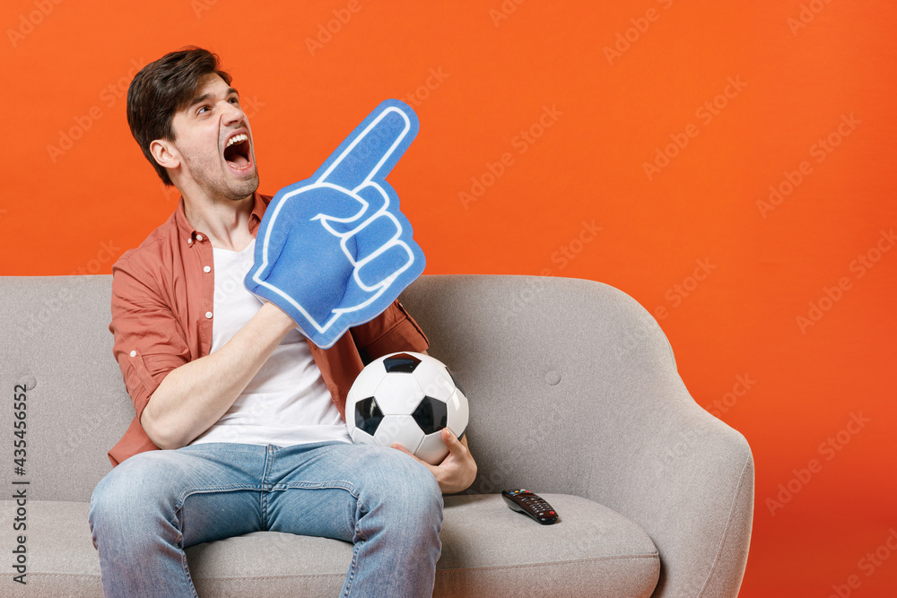 Young man football fan in shirt support team with soccer ball sit sofa home watch tv live stream point foam glove finger aside on workspace isolated on orange background portrait People sport concept.