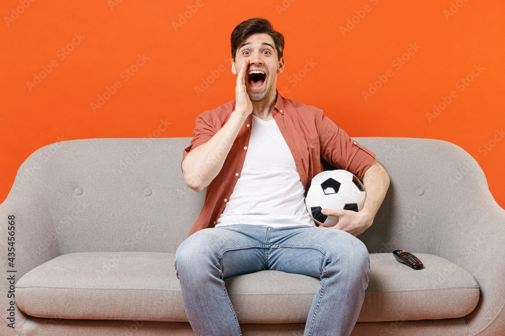 Young exressive fun man football fan in shirt support team with soccer ball sit sofa home watch tv live stream scream news with hands near mouth isolated on orange background. People sport concept
