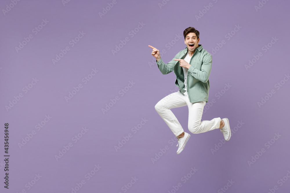 Full length fun excited cool young man in casual mint shirt white t-shirt jump high point index finger aside on copy space area mock up isolated on purple violet background. People lifestyle concept