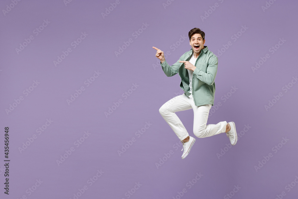 Full length fun excited cool young man in casual mint shirt white t-shirt jump high point index finger aside on copy space area mock up isolated on purple violet background. People lifestyle concept Full length fun excited cool young man in casual mint shirt white t-shirt jump high point index finger aside on copy space area mock up isolated on purple violet background. People lifestyle concept