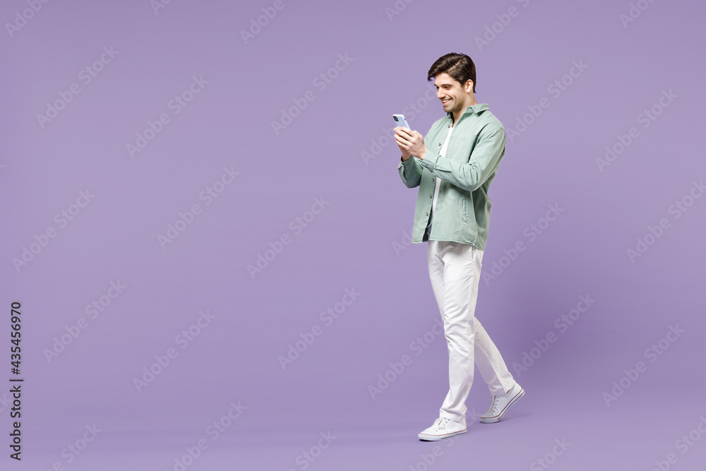 Full length young fun happy man in casual mint shirt white t-shirt using mobile cell phone chat online in social network walk isolated on purple background studio portrait People lifestyle concept. Full length young fun happy man in casual mint shirt white t-shirt using mobile cell phone chat online in social network walk isolated on purple background studio portrait People lifestyle concept.