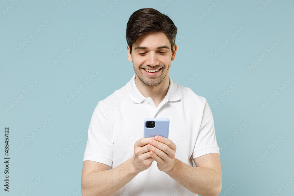 Young fun smiling caucasian man 20s wearing white casual basic t-shirt hold mobile cell phone chat online browsing internet isolated on pastel blue background studio portrait People lifestyle concept. Young fun smiling caucasian man 20s wearing white casual basic t-shirt hold mobile cell phone chat online browsing internet isolated on pastel blue background studio portrait People lifestyle concept.