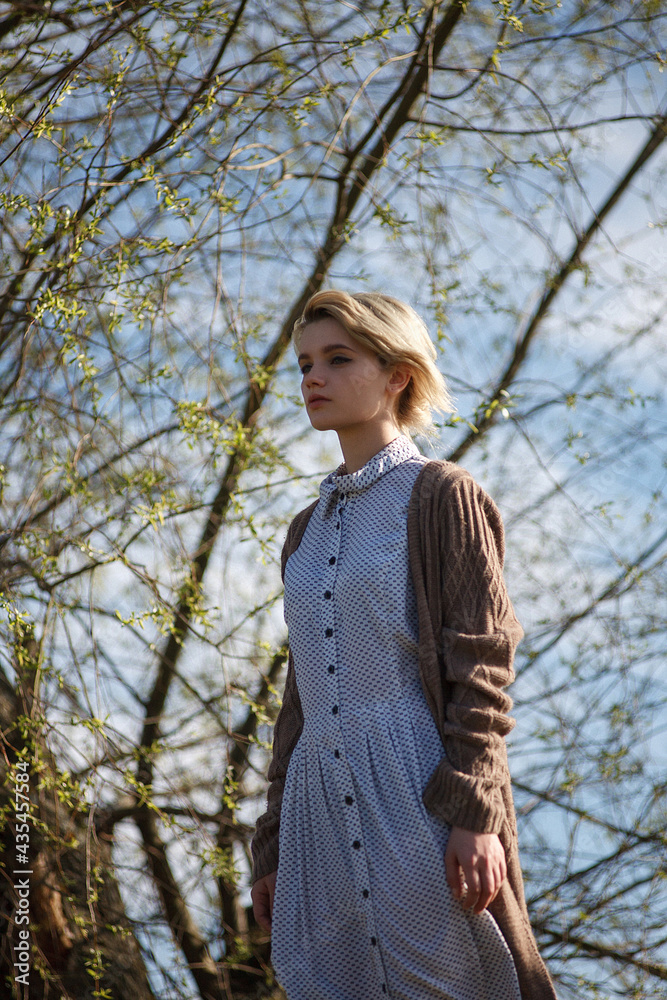 young attractive caucasian female with short hair in spotted dress and sweater standing in front of tree and blue sky on sunny day young attractive caucasian female with short hair in spotted dress and sweater standing in front of tree and blue sky on sunny day
