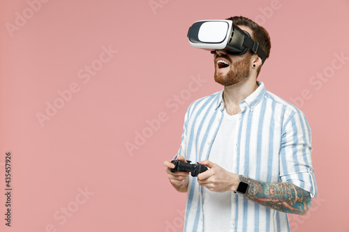 Young excited happy caucasian unshaven man 20s in blue striped shirt playing pc game with joystick console watching in vr headset pc gadget isolated on pastel pink color background studio portrait