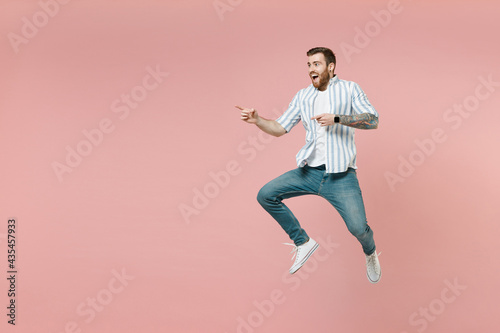 Full length young joyful unshaven man wear blue striped shirt white t-shirt jump high point index finger aside on copy space workspace area isolated on pastel pink background. Tattoo translate fun