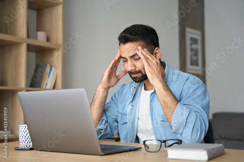 Young indian exhausted business man massaging temples suffering from headache in modern home office with laptop on desk. Overworked burnout academic Hispanic student feeling migraine head strain.