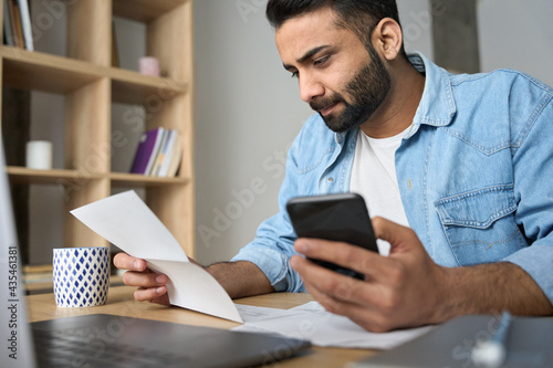 Young indian businessman holding phone reading bank receipt calculating taxes, ethnic man using smartphone mobile application checking bill document, managing money finances, loan expenses.