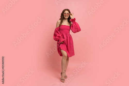Fashion studio portrait of stylish red-haired woman posing in pink lien dress with sleeves over pink background. Full lenght.