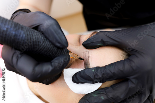 the master of permanent makeup performs a tattoo of the eyebrows with a tattoo machine holding the eye area with a cotton sponge
