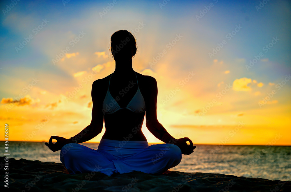 Woman meditating, doing yoga, at the beach, sitting by the seashore, dressed in a white outfit at sunset