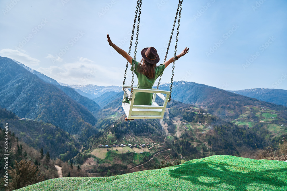 Free happy joyful woman traveler with open arms swinging on chain swing in the mountains, enjoying beautiful view and good life moment