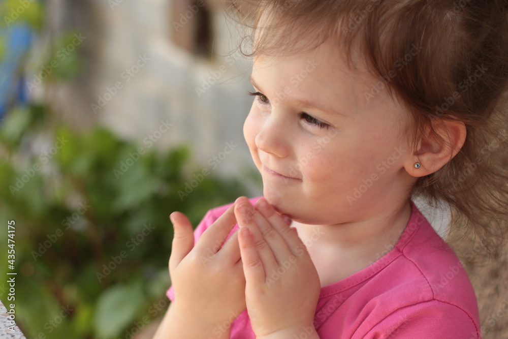 little beautiful smiling red-haired girl in a pink t-shirt sits at a table outdoors with her hands folded little beautiful smiling red-haired girl in a pink t-shirt sits at a table outdoors with her hands folded