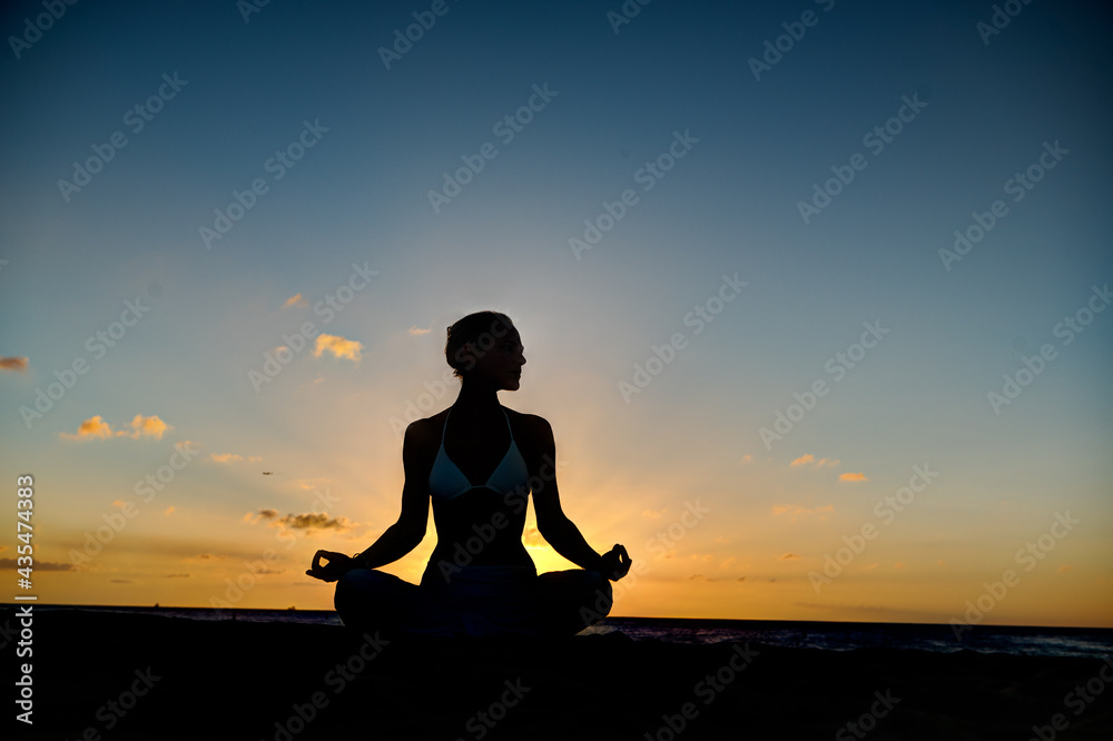 Woman meditating, doing yoga, at the beach, sitting by the seashore, dressed in a white outfit at sunset