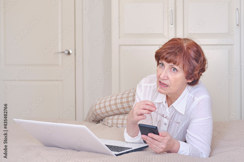 Senior woman wearing glasses, using mobile phone, laptop on the bed at home. Senior woman wearing glasses, using mobile phone, laptop on the bed at home.