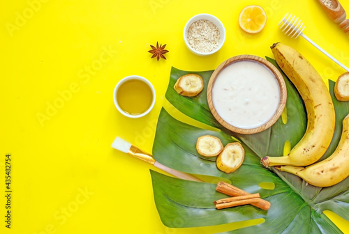 Ingredients for a natural cosmetic facial mask with your own hands. Banana pulp, honey, butter, yolk, oatmeal on a yellow background with exotic leaves, top view, flat lay, copy space. 