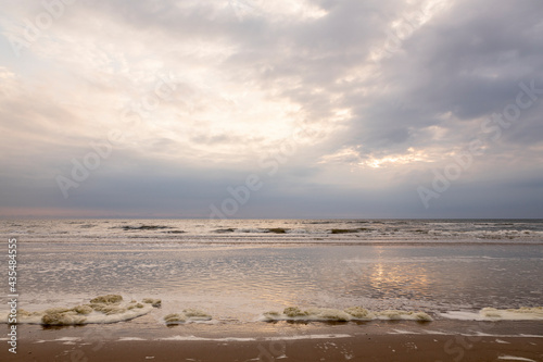 Fototapeta Naklejka Na Ścianę i Meble -  Sea foam washing up on the Dutch North sea beach during sunset on overcast day reflecting in the calm water in the foreground