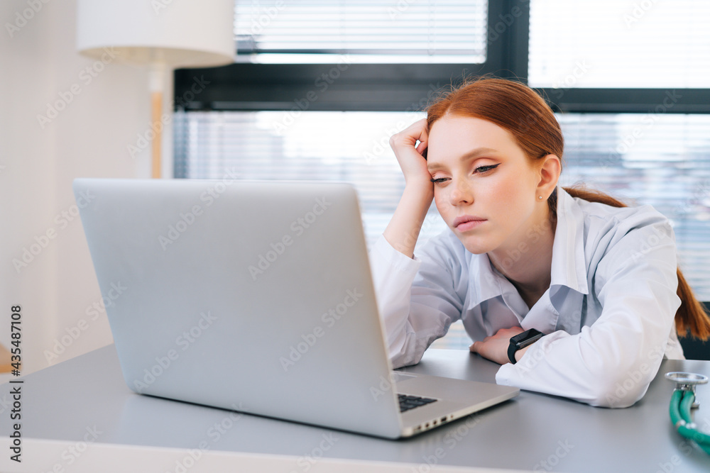 Close-up portrait of tired exhausted young female doctor in white coat sitting at desk with laptop. Front view of overworked woman physician in hospital office on background of window. Close-up portrait of tired exhausted young female doctor in white coat sitting at desk with laptop. Front view of overworked woman physician in hospital office on background of window.