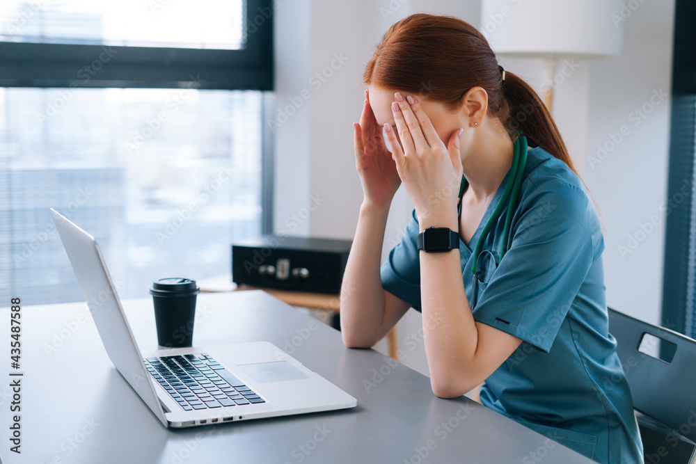 Side view of tired exhausted young female doctor in medical uniform having headache and rubbing head wiht hands, during working on laptop sitting at desk near window in hospital office of medic clinic Side view of tired exhausted young female doctor in medical uniform having headache and rubbing head wiht hands, during working on laptop sitting at desk near window in hospital office of medic clinic