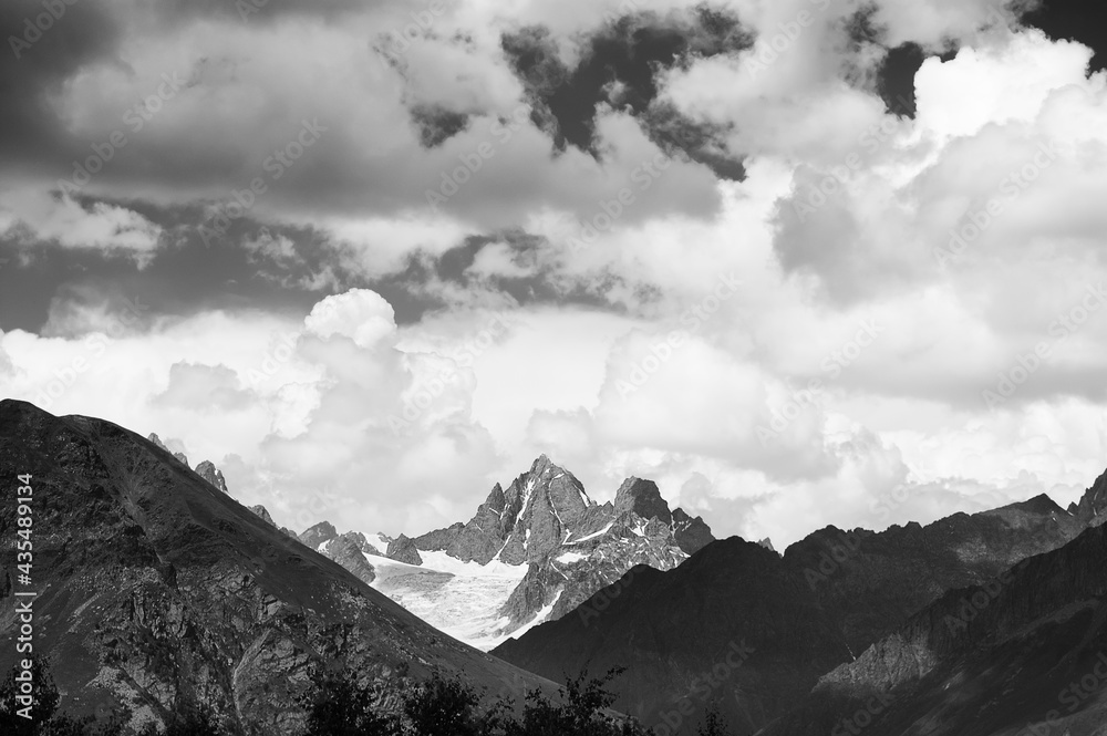 Black and white high rocky mountains with snow and glacier, sunlit sky with clouds at summer