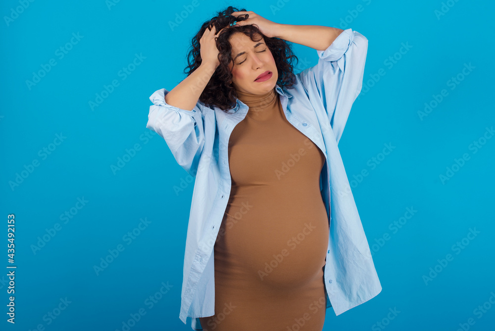 young Arab pregnant woman wearing dress against blue wall suffering from strong headache desperate and stressed because of overwork. Depression and pain concept. young Arab pregnant woman wearing dress against blue wall suffering from strong headache desperate and stressed because of overwork. Depression and pain concept.