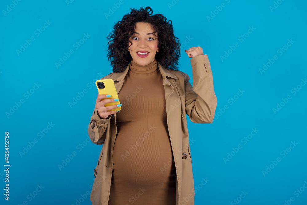 Portrait of young Arab pregnant woman wearing dress against blue wall holding in hands cell celebrating
