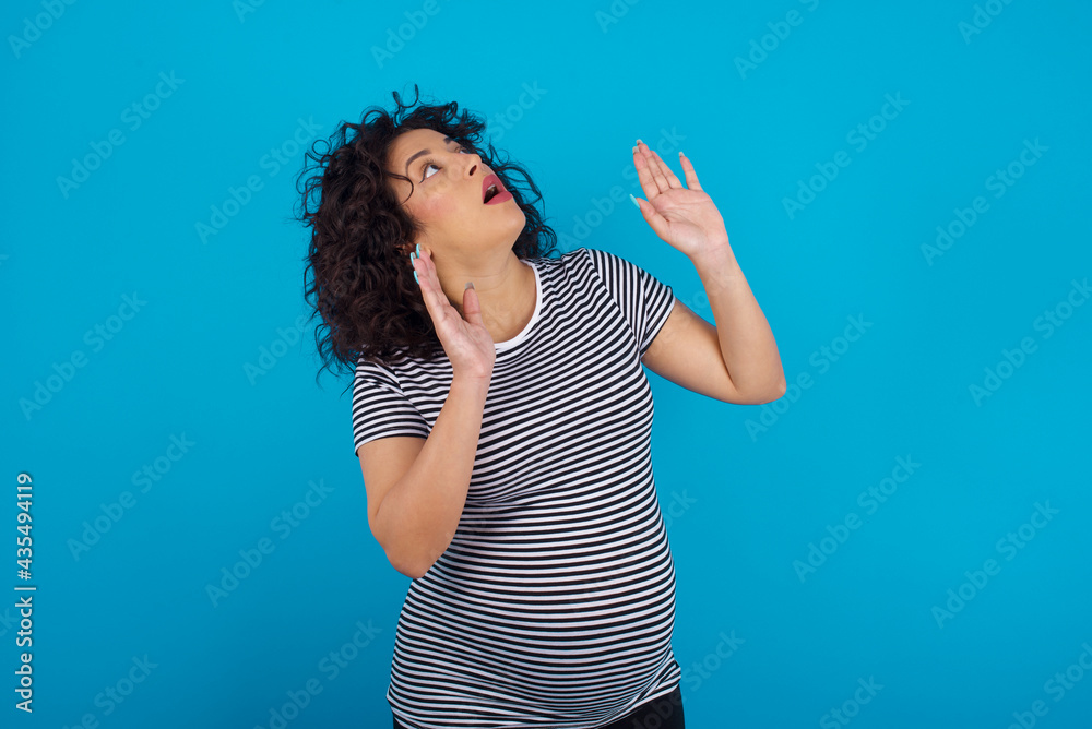 young Arab pregnant woman wearing stripped T-shirt against blue wall keeps palms forward and looks with fright above on ceiling tries to defense herself from invisible danger opens mouth. young Arab pregnant woman wearing stripped T-shirt against blue wall keeps palms forward and looks with fright above on ceiling tries to defense herself from invisible danger opens mouth.