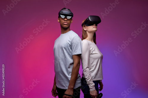 Two calm young man and woman in VR helmets compete in a virtual reality game. Two gamers in virtual reality helmets play VR games, hold controllers. VR gaming, leisure with friends, stand back to back