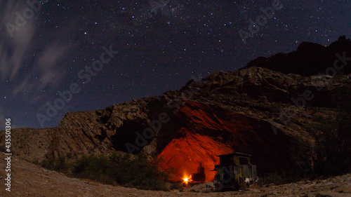 Black shadow of person camping in a cave.   It is night time and stars are visible in the sky. The cave is filled with the orange light of the fire.