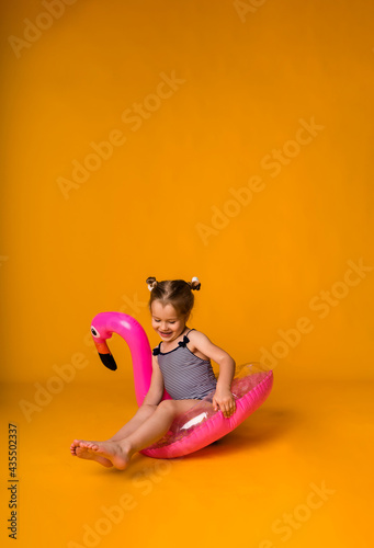 happy little blonde girl in a striped swimsuit sits in a pink inflatable circle with a flamingo on a yellow background with a copy of the space. Summer background