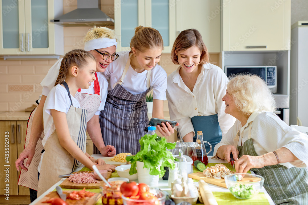 Positive beautiful women use mobile phone while cooking, watching video recipe or tips for cooking, interested in culinary. side view on family dressed in apron, at home in cozy bright kitchen