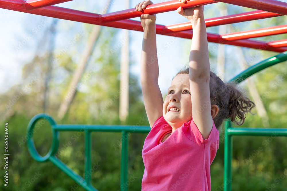 Fototapeta premium child and sport. little active emotional pensive girl on playground in park, hanging on horizontal bar, doing sports exercise outdoors. happy childhood. Kid play on school, kindergarten yard in summer