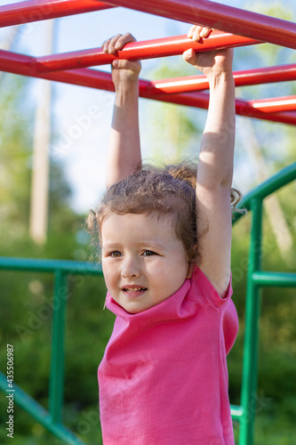 child and sport. little active emotional pensive girl on playground in park, hanging on horizontal bar, doing sports exercise outdoors. happy childhood. Kid play on school, kindergarten yard in summer © Елена Якимова