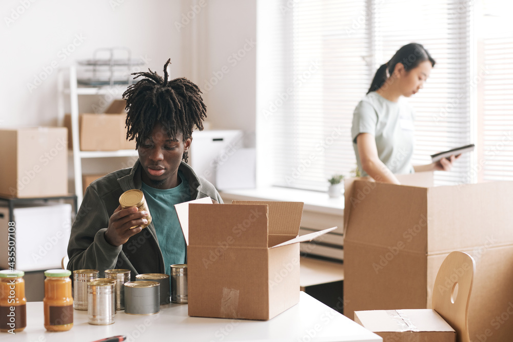 Young African-American man packing canned food to boxes at charity and donations event, copy space Young African-American man packing canned food to boxes at charity and donations event, copy space