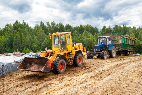 A bulldozer and a tractor compact the cut grass in a silage trench.