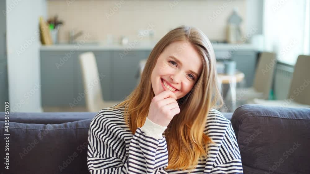 Portrait of beautiful cute young woman in striped sweatshirt smiling sincerely and posing in shy but playful way while looking at camera sitting on sofa at home Portrait of beautiful cute young woman in striped sweatshirt smiling sincerely and posing in shy but playful way while looking at camera sitting on sofa at home